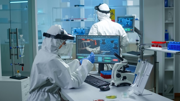 Médico químico en traje de ppe trabajando en pc mientras técnico de laboratorio le trae muestras de sangre