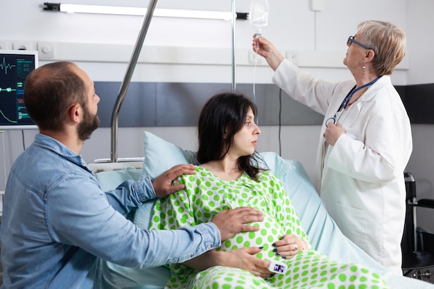 Foto gratuita médico sénior comprobando la bolsa de goteo intravenoso, explicando la cirugía de cesárea a una mujer embarazada en la sala del hospital. mujer con embarazo acostada en la cama discutiendo la maternidad con el futuro padre