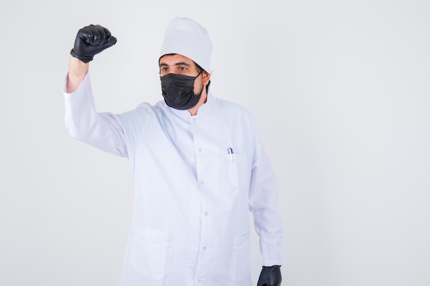 Médico de sexo masculino joven en uniforme blanco mostrando levantando el puño y mirando confiado, vista frontal.