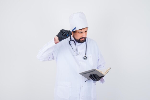 Médico de sexo masculino en uniforme blanco mirando su cuaderno mientras piensa y mira concentrado, vista frontal.