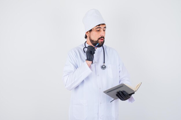 Médico de sexo masculino en uniforme blanco mirando su cuaderno mientras piensa y mira concentrado, vista frontal.