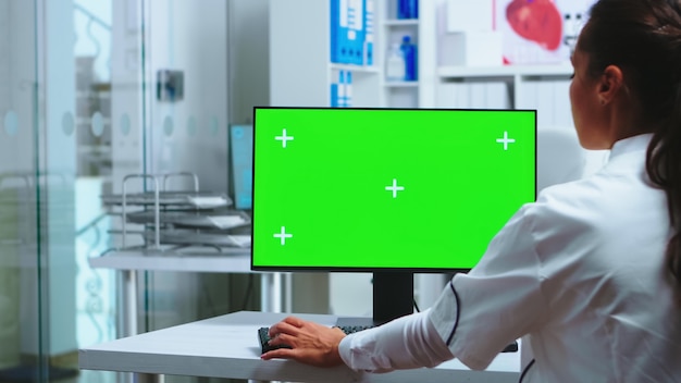 El médico se sienta en la computadora con pantalla verde en blanco en el gabinete del hospital y el asistente en uniforme azul con radiografía. Médico en bata blanca trabajando en el monitor con clave de croma en el gabinete de la clínica para che