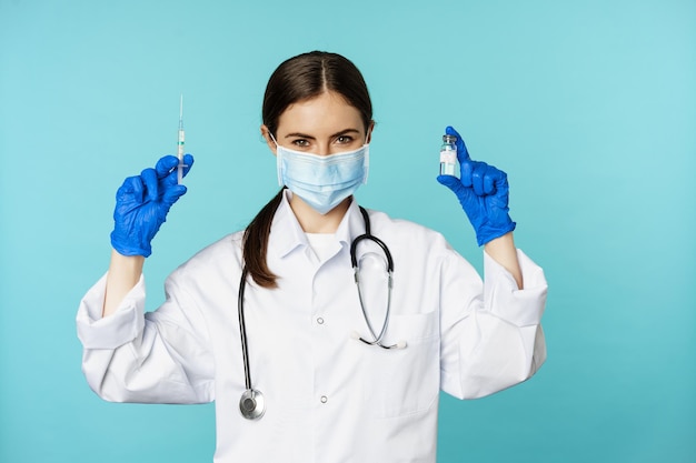 Médico sonriente del personal médico con mascarilla y guantes de goma que muestra jeringa y vacuna de covid p...