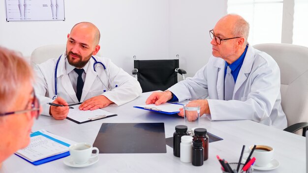 Médico sosteniendo la presentación sobre los síntomas de los pacientes frente a los pasos de investigación de planificación del equipo médico. Equipo médico con conferencia discutiendo sobre enfermedades de personas sentadas en la oficina del hospital