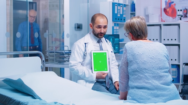 Médico sosteniendo una tableta de pantalla verde con maqueta aislada frente a un paciente anciano en un moderno hospital o clínica privada. Maqueta de croma lista para su aplicación, texto, video u otro activo digital