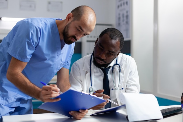 Médico terapeuta afroamericano discutiendo el tratamiento de la salud con un asistente que analiza los síntomas de la enfermedad durante el examen de salud en el consultorio del hospital. Equipo médico que trabaja en la prescripción de medicamentos.