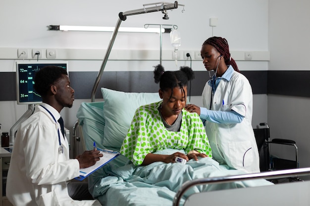 Médico terapeuta afroamericano escuchando los pulmones del paciente con estetoscopio discutiendo la experiencia médica con el terapeuta. Mujer joven enferma descansando en la cama recuperándose después de la cirugía en la sala del hospital