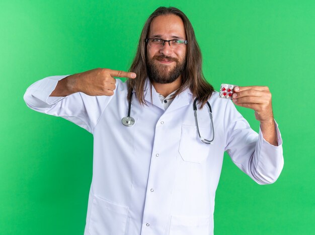 Médico varón adulto complacido con bata médica y estetoscopio con gafas mostrando y apuntando al paquete de cápsulas mirando a cámara aislada en la pared verde