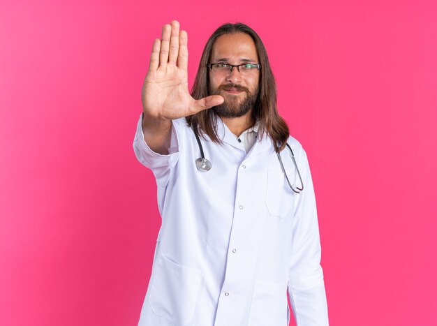Médico varón adulto complacido vistiendo bata médica y estetoscopio con gafas mirando a la cámara haciendo gesto de parada aislado en la pared rosa