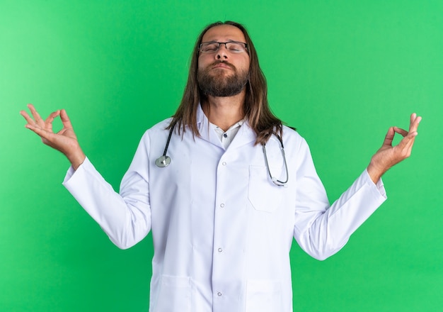 Médico varón adulto pacífico vistiendo bata médica y estetoscopio con gafas meditando con los ojos cerrados aislados en la pared verde