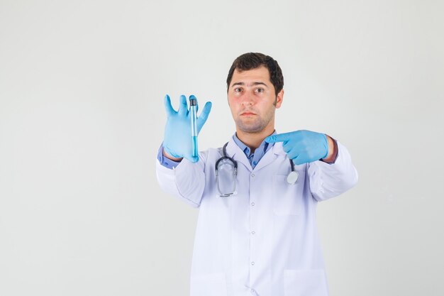 Médico varón apuntando con el dedo al tubo de ensayo en bata blanca, guantes y aspecto serio
