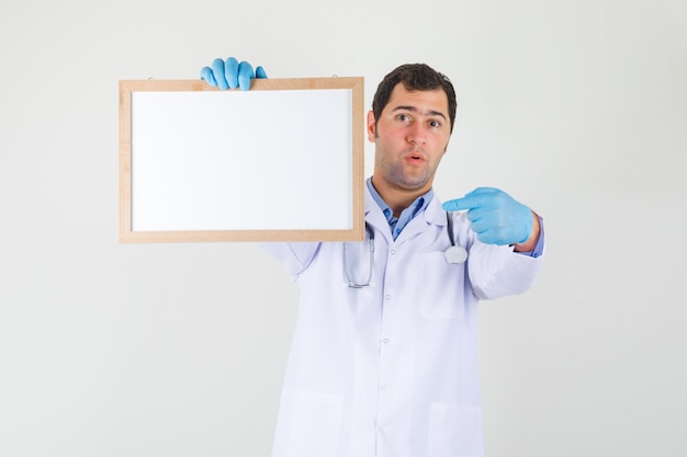 Médico varón apuntando con el dedo a la pizarra blanca en bata blanca, guantes