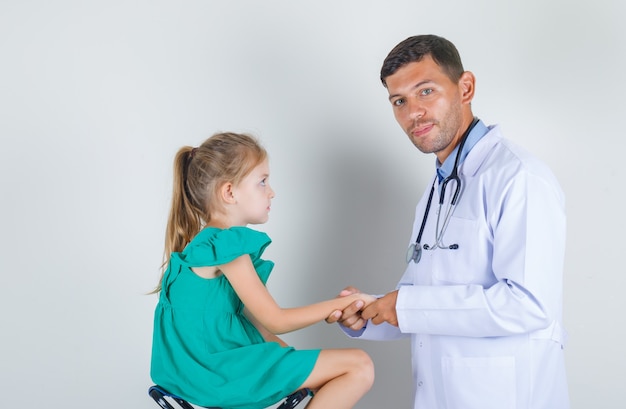 Médico varón auscultando el antebrazo del niño en uniforme blanco en la sala de examen