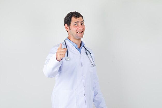 Médico varón en bata blanca apuntando con el dedo a la cámara y mirando alegre