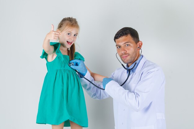 Médico varón en bata blanca escuchando latidos mientras el niño mostrando el pulgar hacia arriba y mirando alegre