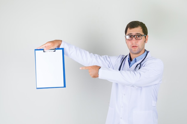 Médico varón en bata blanca, gafas apuntando con el dedo en el portapapeles