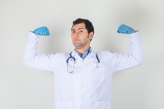 Médico varón en bata blanca, guantes mostrando los músculos levantando las manos y luciendo orgulloso