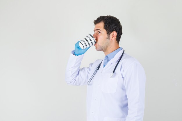 Médico varón bebiendo una taza de café en bata blanca, guantes y mirando sediento.