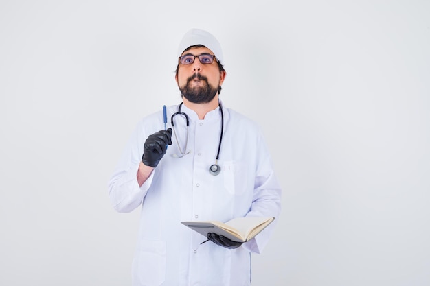 Médico varón escribiendo mientras piensa en uniforme blanco, gafas y mirando concentrado, vista frontal.