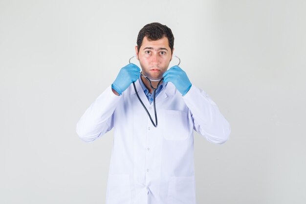 Médico varón con estetoscopio en bata blanca, guantes y mirando enfocado. vista frontal.