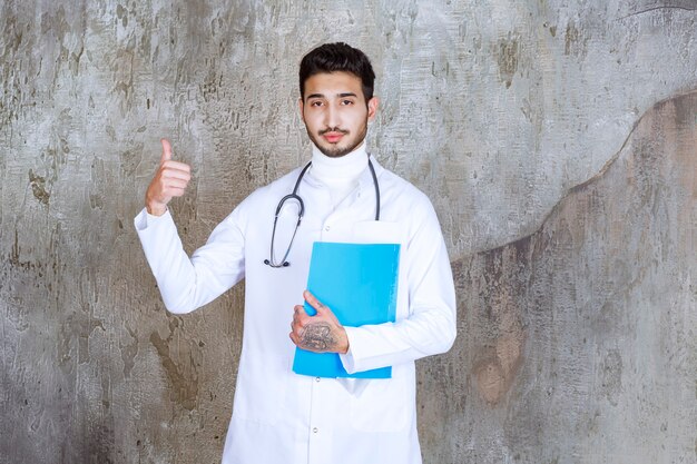 Médico varón con estetoscopio sosteniendo una carpeta azul y mostrando un signo de mano positivo.