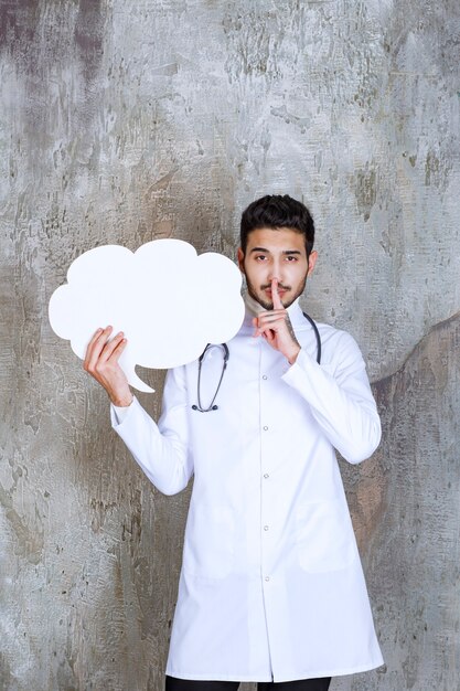 Médico varón con estetoscopio sosteniendo un tablero de información de forma de nube en blanco y pidiendo silencio.