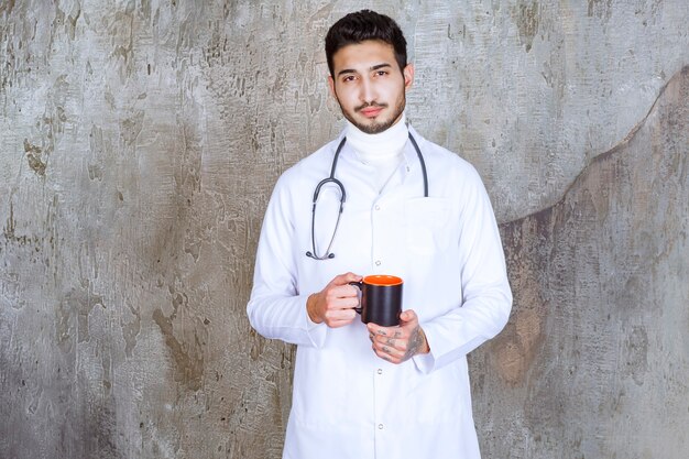 Médico varón con estetoscopio sosteniendo una taza de café y calentando las manos.