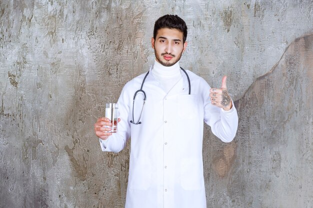 Médico varón con estetoscopio sosteniendo un vaso de agua pura y mostrando un signo de mano positivo.