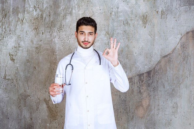 Médico varón con estetoscopio sosteniendo un vaso de agua pura y mostrando un signo de mano positivo