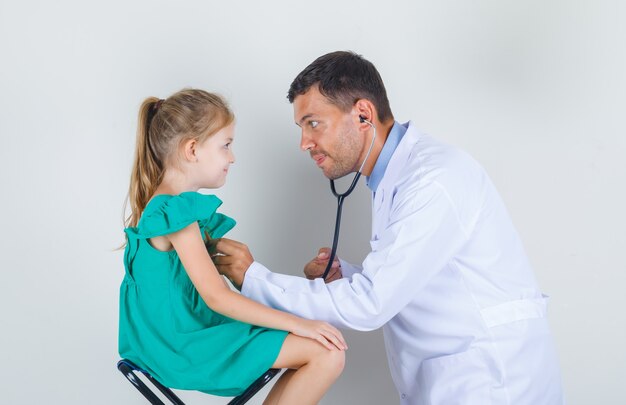 Médico varón examinando el corazón de la niña con estetoscopio en uniforme blanco y mirando contento