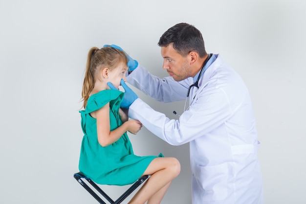 Médico varón examinando los ojos de la niña en uniforme blanco, guantes y mirando con cuidado