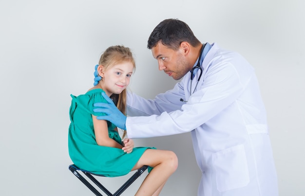 Foto gratuita médico varón examinando las orejas de la niña en uniforme blanco, guantes y mirando con cuidado. vista frontal.