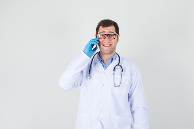 Médico varón hablando por teléfono en bata blanca, guantes, gafas y mirando alegre