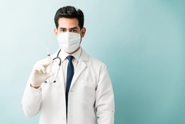 Médico varón hispano sosteniendo una jeringa mientras usa una máscara contra un fondo aislado