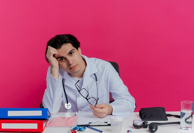 Foto gratuita médico varón joven aburrido con bata médica y estetoscopio sentado en el escritorio con herramientas médicas sosteniendo gafas tocando la cabeza aislada en la pared rosa