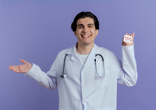Médico varón joven alegre con bata médica y un estetoscopio que muestra el paquete de cápsulas médicas y la mano vacía aislada en la pared púrpura