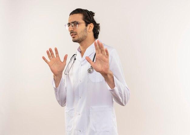 Médico varón joven aprensivo con gafas ópticas vistiendo bata blanca con estetoscopio levantando las manos en la pared blanca aislada con espacio de copia