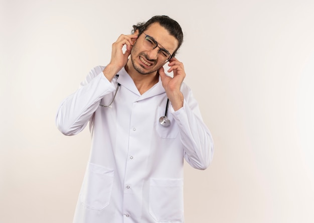 Médico varón joven aprensivo con gafas ópticas vistiendo túnica blanca con estetoscopio orejas cerradas en pared blanca aislada con espacio de copia