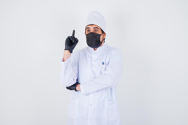 Médico varón joven apuntando hacia arriba en uniforme blanco y mirando vacilante, vista frontal.