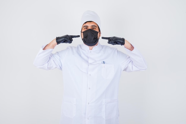 Médico varón joven apuntando a sí mismo en uniforme blanco y mirando orgulloso, vista frontal.