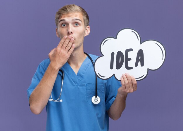 Médico varón joven asustado vistiendo uniforme médico con estetoscopio sosteniendo la boca cubierta de burbuja idea con la mano aislada en la pared azul