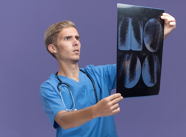 Médico varón joven asustado vistiendo uniforme médico con estetoscopio sosteniendo y mirando rayos x aislado en la pared azul