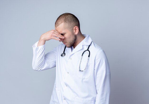 Médico varón joven cansado con bata médica y estetoscopio alrededor de su cuello tapándose la nariz con los ojos cerrados aislado sobre fondo blanco con espacio de copia