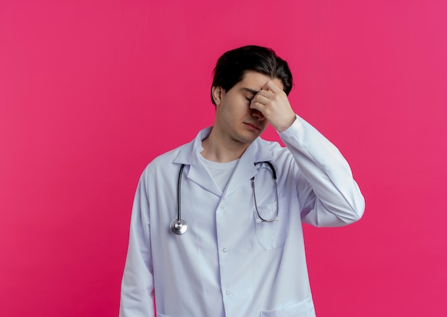 Médico varón joven cansado con bata médica y estetoscopio sosteniendo la nariz con los ojos cerrados aislado en la pared rosa con espacio de copia