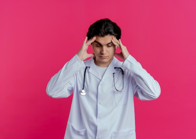 Médico varón joven cansado con bata médica y estetoscopio tocando la cabeza con los ojos cerrados aislado en la pared rosa con espacio de copia