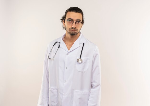 Médico varón joven cansado con gafas ópticas vistiendo bata blanca con estetoscopio en pared blanca aislada con espacio de copia