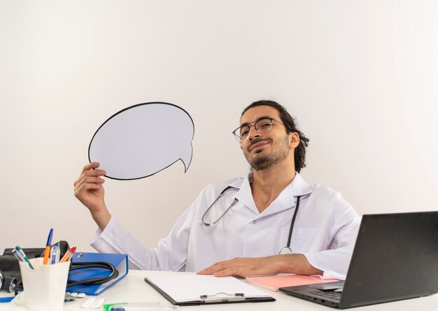 Médico varón joven complacido con gafas médicas vistiendo bata médica con estetoscopio