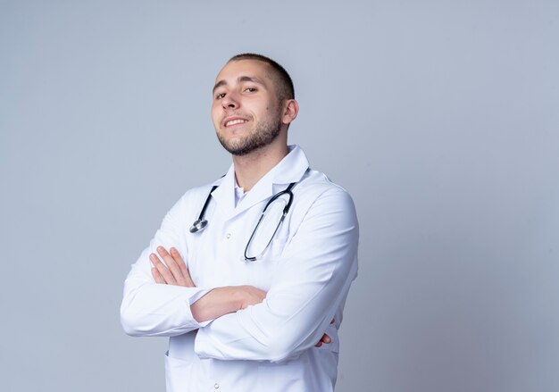 Médico varón joven confiado con bata médica y un estetoscopio alrededor de su cuello de pie con postura cerrada y sonriendo aislado en blanco con espacio de copia