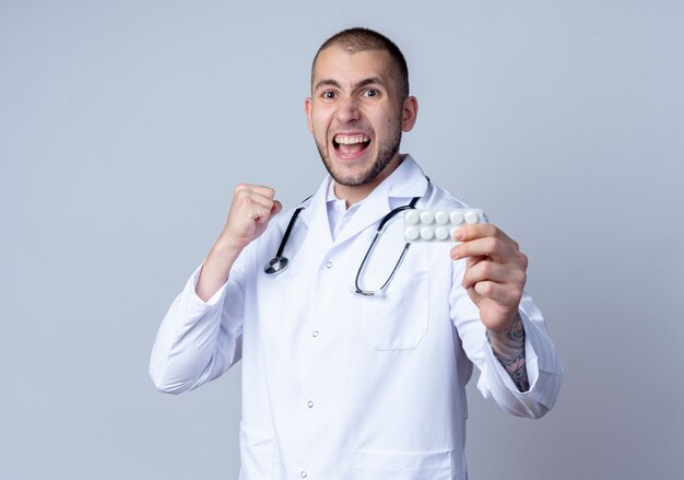Médico varón joven confiado con bata médica y un estetoscopio alrededor de su cuello sosteniendo un paquete de tabletas médicas y apretando el puño aislado en blanco