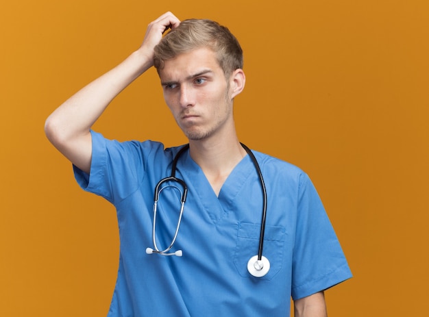 Médico varón joven confundido vistiendo uniforme médico con estetoscopio rascándose la cabeza aislada en la pared naranja